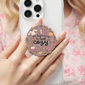 May include: A circular phone grip featuring a light brown background with the text "it's beginning to look cozy" in black script. The design includes small illustrations of a sweater, mug, pumpkin, candle, beanie, scarf, leaves, and mushrooms.
