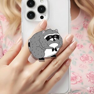 May include: A clear smartphone case featuring a circular phone grip attachment. The grip displays a cartoon illustration of a round, grey raccoon with a white belly, black eye mask, and small black paws, centered on the back of the mobile device.