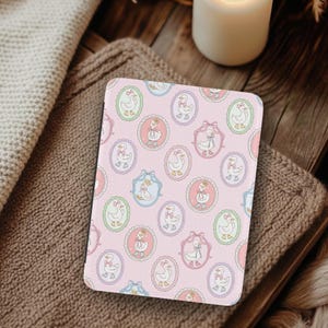 Puede incluir: Una funda rectangular rosa con un patrón repetido de gansos de dibujos animados en marcos ovalados. Los gansos llevan lazos y vestidos. Los marcos son de colores pastel, incluyendo rosa, azul y verde. La funda está sobre una superficie marrón.