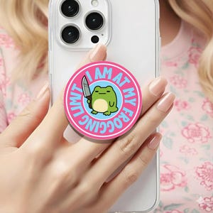May include: A pink phone grip with the text "I AM AT MY FROGGING LIMIT" encircling a cartoon frog holding a knife. The frog is green with a light blue background. The phone grip is attached to a clear phone case.