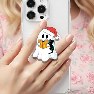 May include: A white ghost-shaped phone grip with a red Santa hat, holding an orange book with a black cat. The phone accessory is attached to a clear phone case. The design is festive and whimsical.