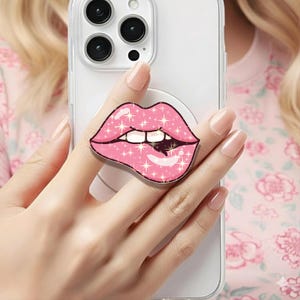 Puede incluir: Un soporte de teléfono emergente rosa con un diseño de labios brillantes. Los labios tienen dientes blancos y un interior oscuro, con detalles de purpurina en forma de estrella. El soporte está adherido a una funda transparente para teléfono.