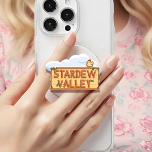 May include: A phone grip with the words "Stardew Valley" on a wooden sign design, with a cloud and chicken graphic. The grip is attached to a clear phone case.