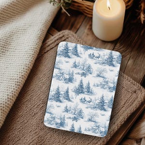 May include: A rectangular coaster with a blue and white winter scene design featuring houses, trees, and a horse-drawn carriage. The coaster is resting on a brown knitted surface, with a lit candle and a beige knitted blanket in the background.