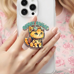 Pode incluir: Um suporte de telefone com um gato tigrado laranja de desenho animado usando óculos e uma mochila. O texto "QUEST SQUAD" está acima do gato. O suporte está preso a uma capa de telefone transparente.