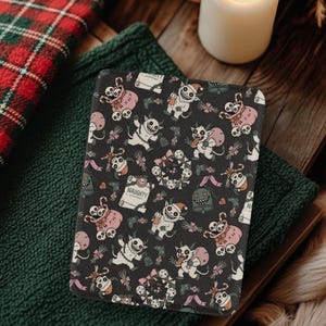 Könnte beinhalten: Ein dunkelgraues, gemustertes Objekt mit Cartoon-Monstern, Schleifen und dem Wort "NAUGHTY". Das Design enthält rosa, weiße und schwarze Elemente. Es liegt auf einer Holzoberfläche mit einem grünen Tuch und einem rot-grünen Karostoff.