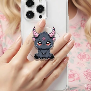 May include: A phone grip featuring a cartoonish, dark gray goat with pink horns and a pink star on its forehead. The goat has large red eyes and is sitting down. The phone grip is attached to a clear phone case.