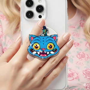 May include: A blue cat phone grip with a cartoon design. The cat has large yellow eyes, a wide grin, and a small hat. The grip is attached to a clear phone case. The cat's outline and the grip's edge have a sparkly, glittery effect.