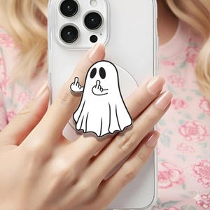 May include: A white ghost-shaped phone grip with black eyes and a black outline. The ghost is giving the middle finger with both hands. The phone grip is attached to a clear phone case.
