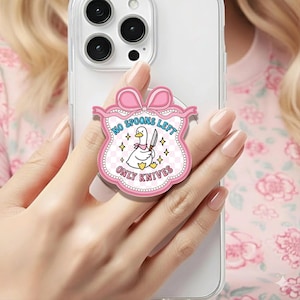 May include: A pink phone grip with a cartoon duck holding a knife. The grip has the text "NO SPOONS LEFT ONLY KNIVES" in a decorative banner shape with a bow at the top. The grip is attached to a white phone.