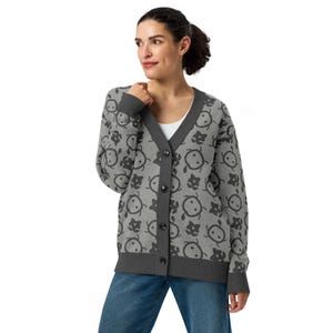 Könnte beinhalten: Graue Strickjacke mit einem sich wiederholenden Muster aus schwarzen Cartoon-Gesichtern und floralen Formen. Die Strickjacke hat einen V-Ausschnitt, dunkelgraue Paspeln und passende Knöpfe. Die Ärmel und der Saum sind ebenfalls dunkelgrau eingefasst.