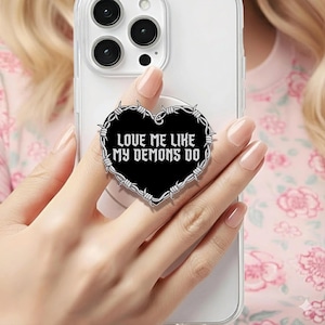 May include: A black heart-shaped phone grip with the text "LOVE ME LIKE MY DEMONS DO" in white. The heart is outlined with a barbed wire design. The phone grip is attached to a clear phone case.