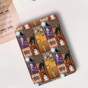 Peut inclure: Une housse de tablette sur le thème d'Halloween avec un motif patchwork. La housse présente des carrés avec des images de fantômes, de chats noirs, de squelettes et le mot "BOO". La palette de couleurs comprend l'orange, le noir, le violet et le blanc.