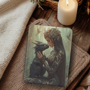 Könnte beinhalten: Ein dekorativer Artikel mit Fantasy-Thema, der eine Frau und einen Drachen zeigt. Die Frau mit langen dunklen Haaren trägt ein goldverziertes Kleid und einen Blattkranz und berührt das Gesicht des Drachen. Der Drache ist schwarz mit gelben Augen. Der Hintergrund ist ein sanftes Grün.
