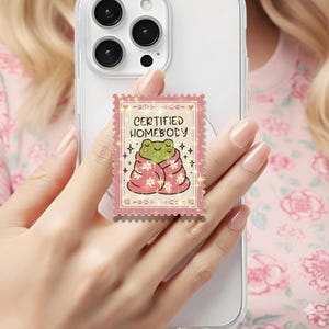 May include: A pink phone grip with a cartoon frog in a blanket. The grip, resembling a stamp, has the text "CERTIFIED HOMEBODY". The frog is green, and the blanket is pink with white flower details. The grip is attached to a phone.