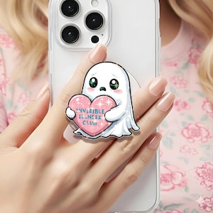 May include: A white ghost-shaped phone grip with a sad expression, holding a pink heart with the text "Invisible Illness Club." The phone grip is attached to a clear phone case.