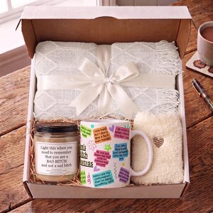 May include: A gift box containing a white knit throw blanket tied with a satin ribbon, a glass jar candle with a label reading 'Light this when you need to remember that you are a BAD BITCH and not a sad bitch', and a white mug with colorful text.