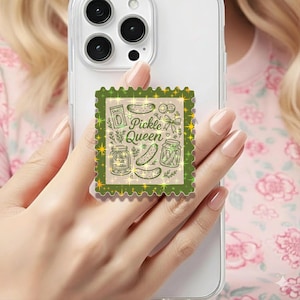 May include: A green and gold phone grip featuring a "Pickle Queen" design. The design includes illustrations of pickles, jars, and the word "Snack". The phone grip is attached to a white phone.