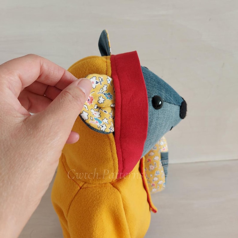 Puede incluir: Un juguete de peluche amarillo con capucha con detalles de denim y tela floral. El juguete tiene un ribete rojo alrededor de la capucha y una bufanda con estampado floral.
