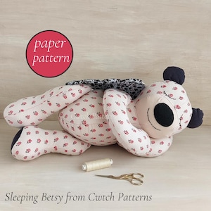 May include: A stuffed bear toy made of cream fabric with a small red floral print, featuring black ears, a black nose, and embroidered closed eyes. A black and white patterned fabric is on the back. Includes a spool of thread and scissors.