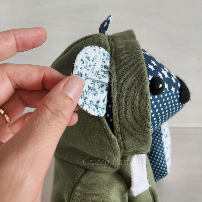 Puede incluir: Un juguete de peluche verde con capucha y un cuerpo estampado en azul y blanco. La capucha tiene un detalle de oreja con estampado floral. El juguete tiene un ojo de bot&oacute;n negro y una nariz negra.