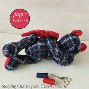 May include: A plush bear toy made of navy blue, white, and red plaid fabric with red accents on the ears and wings. The bear is in a sleeping position. Below the bear are two spools of thread and a pair of metal scissors. Text reads paper pattern.