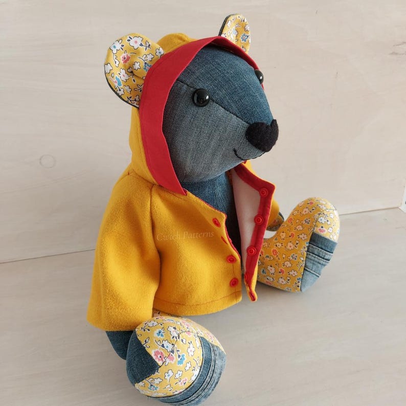 Puede incluir: Un oso de peluche de mezclilla con un impermeable amarillo con capucha roja y tela de estampado floral en las orejas y los pies.