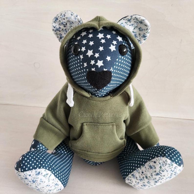 Puede incluir: Un oso de peluche que lleva una sudadera con capucha verde con el texto "Cwtch Patterns" en la parte delantera. El oso tiene un cuerpo estampado en azul y blanco con estrellas blancas en la capucha.