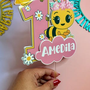May include: A pink and yellow number one cake topper with a cartoon bee wearing a flower crown and the name "AMEDila". The topper has daisy accents and a smiling sun. The background is pink.