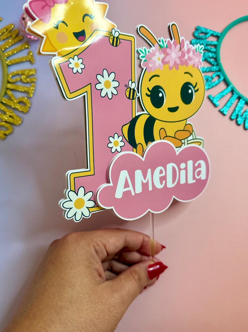 May include: A pink and yellow number one cake topper with a cartoon bee wearing a flower crown and the name "AMEDila". The topper has daisy accents and a smiling sun. The background is pink.