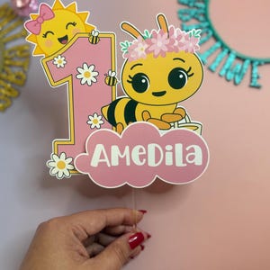May include: A pink and yellow number one cake topper featuring a cartoon bee with a flower crown and a smiling sun with a bow. The name "AMEDILA" is written on a pink cloud. Daisies and bees decorate the number one.