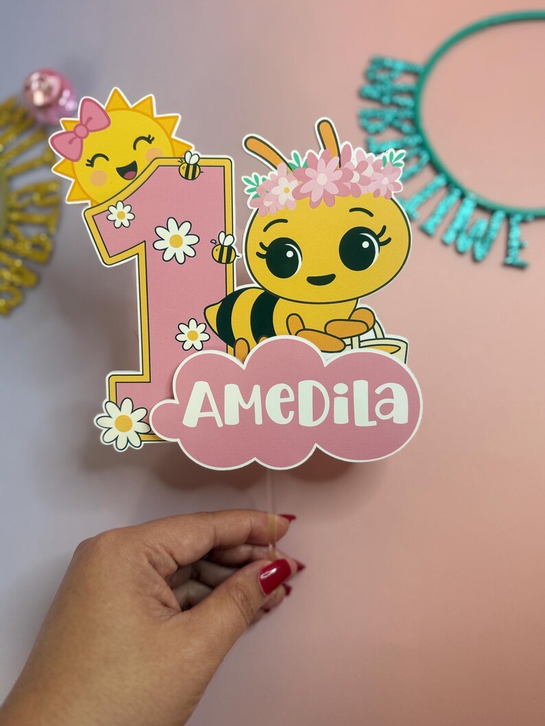 May include: A pink and yellow number one cake topper featuring a cartoon bee with a flower crown and a smiling sun with a bow. The name "AMEDILA" is written on a pink cloud. Daisies and bees decorate the number one.