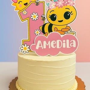 May include: A yellow frosted cake with a decorative topper for a first birthday. The topper features a cartoon bee, a smiling sun, the number 1, and the name "AMEDILA" in pink. The cake sits on a gold scalloped cake board.