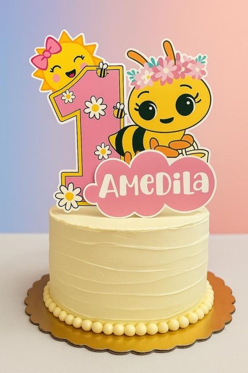 May include: A yellow frosted cake with a decorative topper for a first birthday. The topper features a cartoon bee, a smiling sun, the number 1, and the name "AMEDILA" in pink. The cake sits on a gold scalloped cake board.