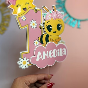 May include: A pink and yellow birthday cake topper featuring a cartoon bee with a flower crown and the number one. The topper includes a smiling sun, daisies, and the name "AMeDila" in white script.