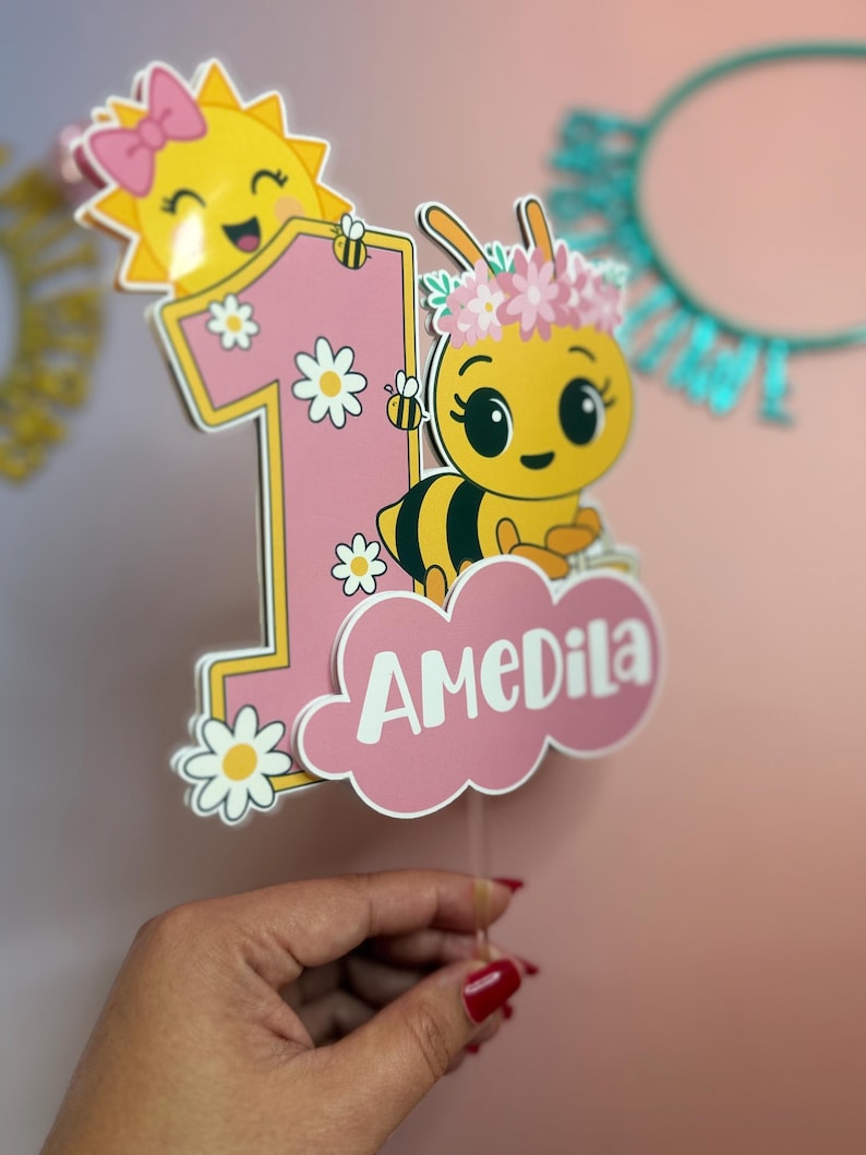 May include: A pink and yellow birthday cake topper featuring a cartoon bee with a flower crown and the number one. The topper includes a smiling sun, daisies, and the name "AMeDila" in white script.