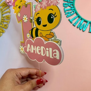 May include: A pink and yellow birthday cake topper featuring a cartoon bee with a flower crown, the number 1, and the name "AMEDILA". The bee is smiling, and the sun is also smiling. The topper is held by a clear plastic stick.