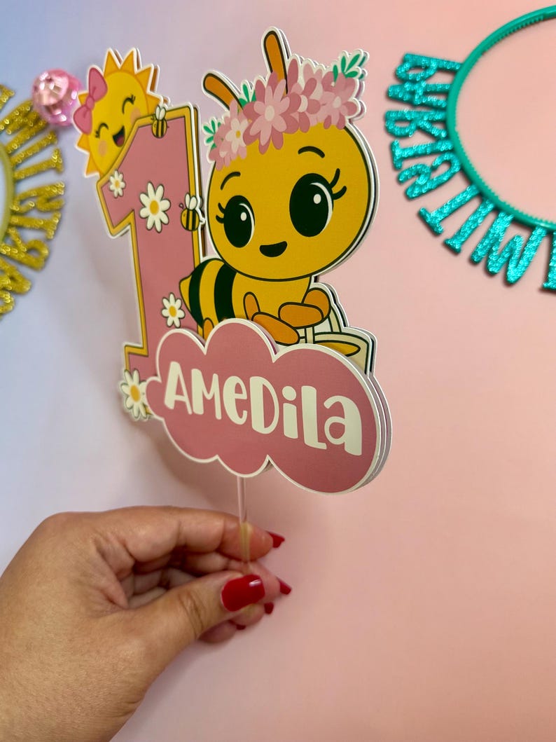 May include: A pink and yellow birthday cake topper featuring a cartoon bee with a flower crown, the number 1, and the name "AMEDILA". The bee is smiling, and the sun is also smiling. The topper is held by a clear plastic stick.