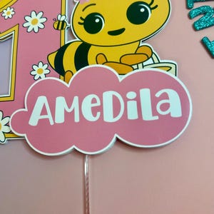 May include: A pink and yellow cake topper featuring a cartoon bee with a flower crown and the name "AMEDILA" in white. The number "1" is also present, along with a smiling sun and daisies. The topper is designed for a birthday celebration.
