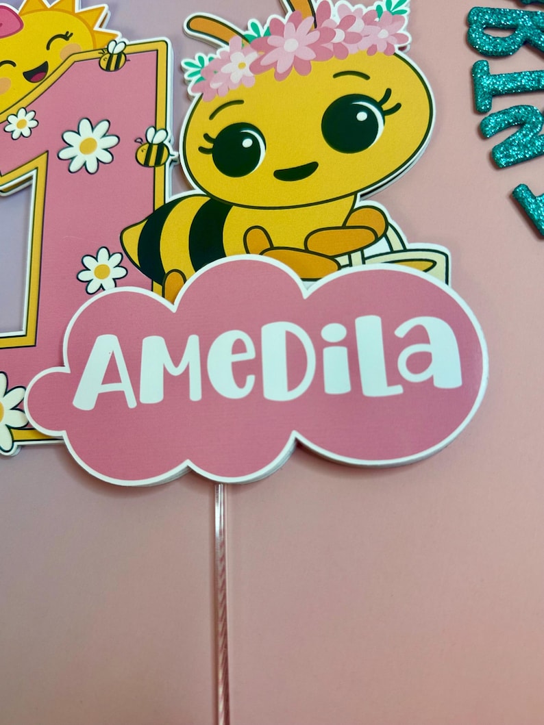 May include: A pink and yellow cake topper featuring a cartoon bee with a flower crown and the name "AMEDILA" in white. The number "1" is also present, along with a smiling sun and daisies. The topper is designed for a birthday celebration.