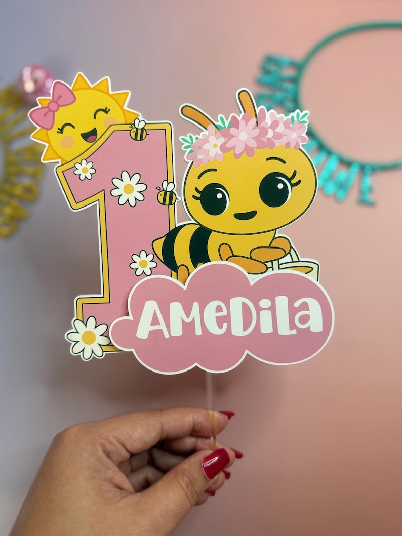May include: A pink and yellow birthday cake topper featuring a cartoon bee with a flower crown, a smiling sun, and the number one. The name "AMEDILA" is written on a pink cloud. Daisies and bees decorate the number one.