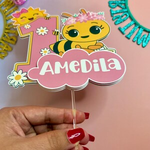 May include: A pink and white cake topper with a cartoon bee, the number 1, and the name "AMeDiLa". The bee is yellow and black, wearing a flower crown. The number 1 has a smiling sun and daisies. The topper is held by a clear stick.