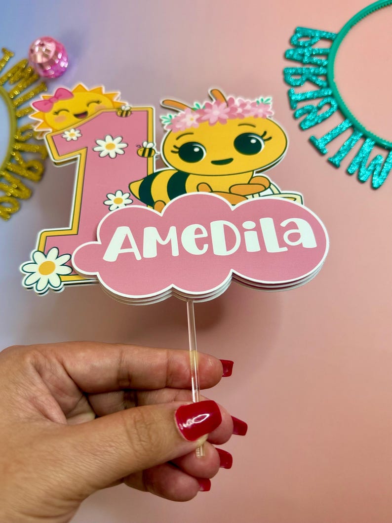 May include: A pink and white cake topper with a cartoon bee, the number 1, and the name "AMeDiLa". The bee is yellow and black, wearing a flower crown. The number 1 has a smiling sun and daisies. The topper is held by a clear stick.