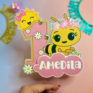 May include: A pink and yellow birthday cake topper featuring a cartoon bee wearing a flower crown and a smiling sun. The number "1" is decorated with daisies and bees, with the name "AMEDILA" on a pink cloud.