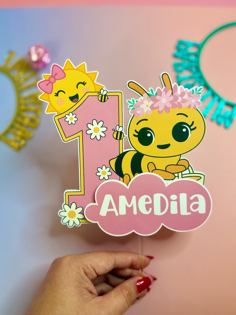 May include: A pink and yellow birthday cake topper featuring a cartoon bee wearing a flower crown and a smiling sun. The number "1" is decorated with daisies and bees, with the name "AMEDILA" on a pink cloud.