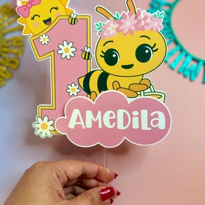 May include: A pink and yellow birthday cake topper featuring a cartoon bee wearing a flower crown and a smiling sun with a bow. The number one is pink with daisy accents, and the name "AMEDILA" is displayed on a pink cloud.