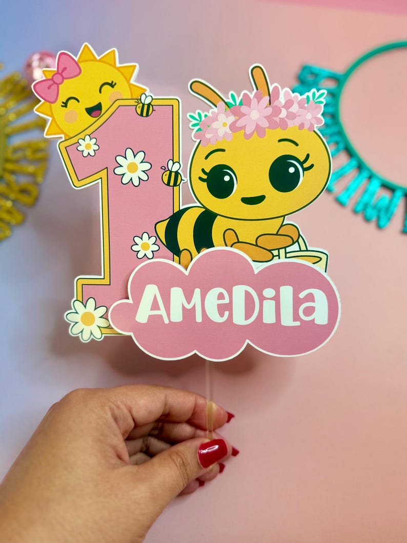 May include: A pink and yellow birthday cake topper featuring a cartoon bee wearing a flower crown and a smiling sun with a bow. The number one is pink with daisy accents, and the name "AMEDILA" is displayed on a pink cloud.