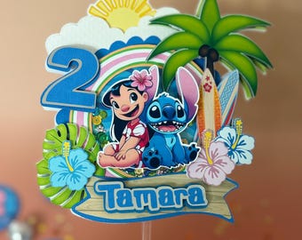 May include: A colorful cake topper featuring the characters Lilo and Stitch, a blue number 2, and the name "Tamara". The design includes a rainbow, palm tree, surfboards, and tropical flowers, perfect for a birthday celebration.
