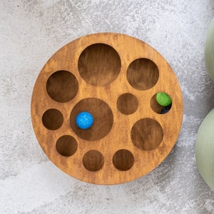 May include: A wooden circular tray with multiple circular cutouts of varying sizes. The tray has a light brown finish and contains two small balls, one blue and one green.