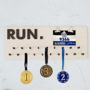 May include: A wooden medal display with the word "RUN." printed in black. The display has 24 hooks for hanging medals. Three medals are hanging on the hooks. The medals are gold, bronze, and blue. A blue and black marathon race bib is attached to the display with two silver clips. The bib reads "adidas SIMON 9366 124th Boston Marathon Virtual Experience John Hancock."